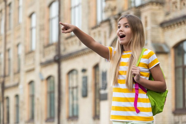 Ragazza teenager sorpresa che punta il dito in avanti in piedi a scuola sfocata all'aperto copia spazio