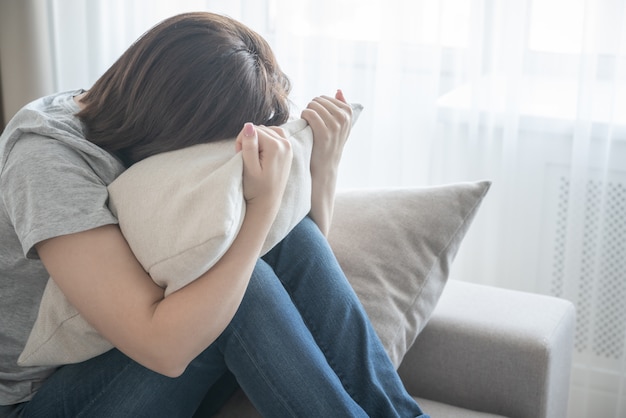 Ragazza triste che si siede sullo strato e che abbraccia un concetto del cuscino, di solitudine e di tristezza