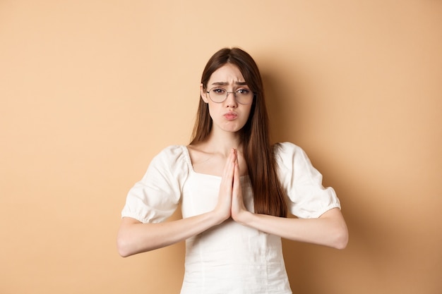 Ragazza triste con gli occhiali che chiede aiuto, per favore e sembra carina, ha bisogno di favore, in piedi sul beige.