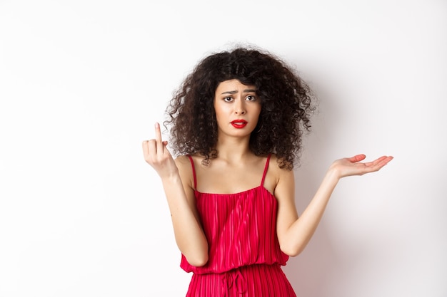 Ragazza triste con i capelli ricci, che mostra il dito senza anello e sembra confusa, si lamenta del fidanzato indeciso, vuole sposarsi, in piedi su sfondo bianco.