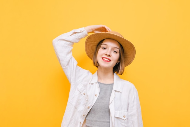 Ragazza turistica sorridente con cappello da sole e abiti casual leggeri che posano alla telecamera su sfondo giallo