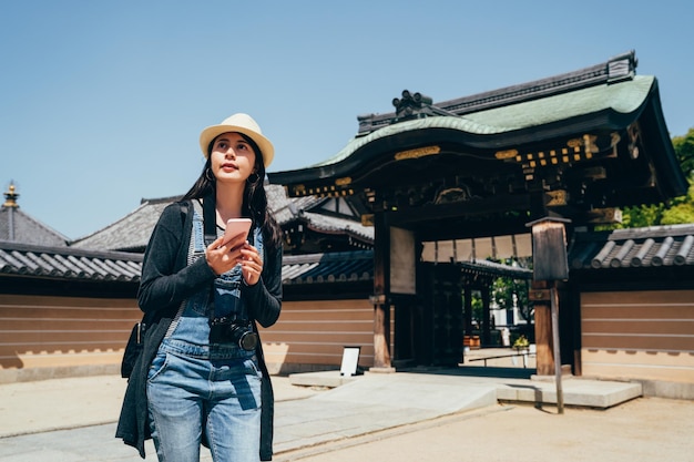 ragazza turistica tenendo il telefono cellulare in piedi all'aperto con cielo blu in una giornata di sole. donna di viaggio che utilizza la guida online sullo smartphone per trovare la via del ritorno in hotel. grande cancello d'ingresso di shitennoji.