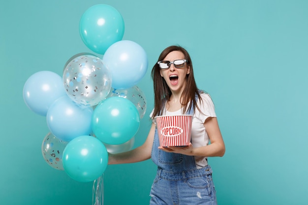 Ragazza urlante con occhiali 3d che guarda un film tiene un secchio di popcorn che celebra con palloncini colorati isolati su sfondo blu turchese. Emozioni di festa di compleanno nel concetto di cinema.