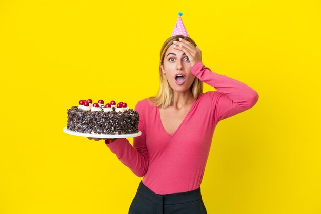 Ragazza uruguaiana bionda che tiene la torta di compleanno isolata su fondo giallo con l'espressione di sorpresa