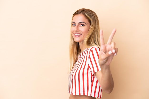 Ragazza uruguaiana bionda isolata su fondo beige che sorride e che mostra il segno di vittoria