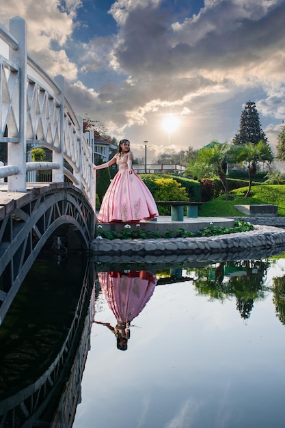 Ragazza vestita da principessa in posa accanto a un ponte e un lago, il suo riflesso è nell'acqua.