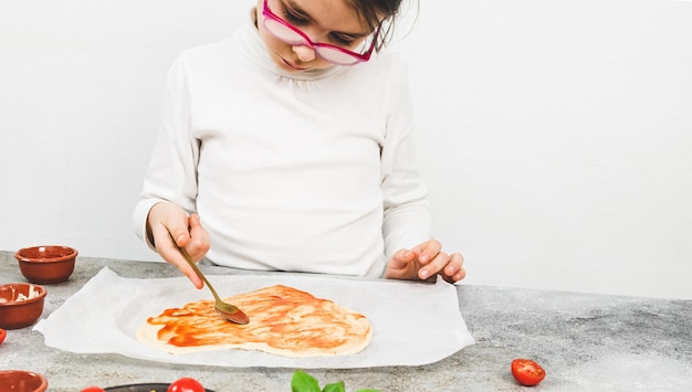 Ragazze caucasiche in un dolcevita bianco spalmano un cucchiaio con salsa di pomodoro su un impasto per pizza a forma di cuore