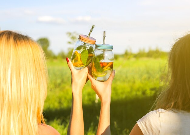 Ragazze che tengono limonata fresca in barattoli con cannucce. Festa estiva hipster con bevande. Stile di vita vegano sano. Ecologico nella natura. Limoni, arance e frutti di bosco con menta nel bicchiere.