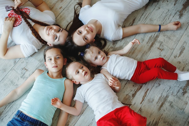 Ragazze con le trecce e un fratello minore, una famiglia in magliette bianche e jeans giacciono sul pavimento testa a testa, sorridono e ridono. Il concetto di una famiglia felice.