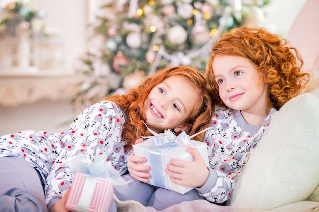 Ragazze dai capelli rossi in pigiama giacciono sul letto contro un albero di Natale con i regali in mano.