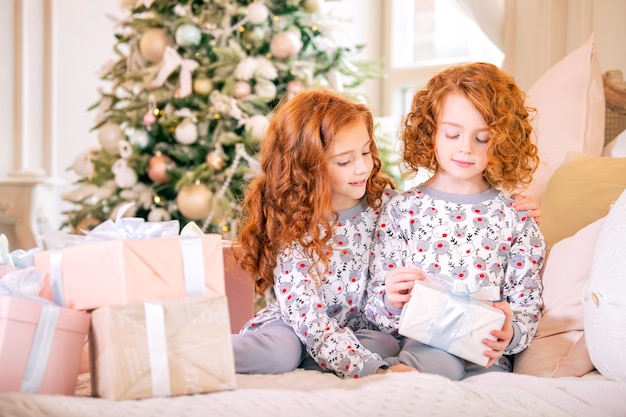 Ragazze dai capelli rossi in pigiama sul letto stanno guardando scatole di regali di Natale contro l'albero di Natale.