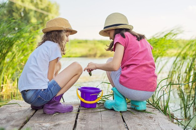 Ragazze dei bambini che giocano insieme sul lago, seduti sul molo di legno, catturando lumache d'acqua nel secchio. Infanzia, estate, natura, concetto di bambini