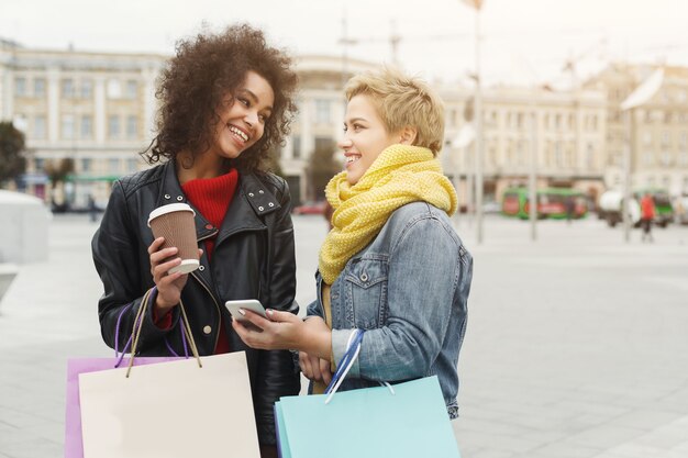 Ragazze felici con borse della spesa e caffè da asporto all'aperto. Amiche sorridenti in abiti casual caldi e luminosi che fanno una passeggiata in città. Amicizia, stile di vita urbano e concetto di svago