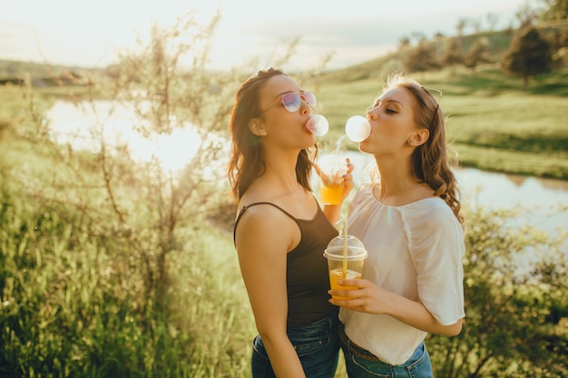 Ragazze felici divertendosi, gonfia di gomma da masticare, bevendo cocktail in bicchiere di plastica con paglia in occhiali da sole, al tramonto, espressione facciale positiva, all'aperto