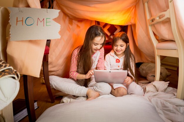Ragazze felici sedute in casa fatta di coperte e che usano tablet digitale