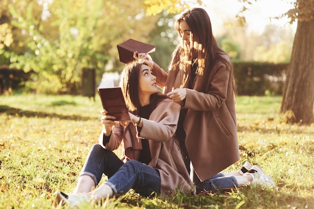 Ragazze gemelle castane che si siedono sull'erba con i libri marroni nelle mani e si guardano, quando uno di loro è in piedi sulle sue ginocchia vicino alla parte posteriore della sorella nel parco di autunno su sfondo sfocato.