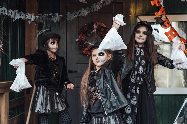 Ragazze spaventose sorelle amici che celebrano halloween Giocando a treak or treat game sulla veranda con ghirlanda Borse con dolci in manoTrucco terrificante teschio viso Costumi eleganti da strega Festa per bambini