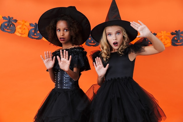 ragazze streghe spaventate in costumi di halloween neri che guardano la telecamera e mostrano il gesto di arresto isolato su un muro di zucca arancione