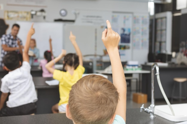 Ragazzi delle scuole che alzano la mano in classe a scuola con l'insegnante sullo sfondo