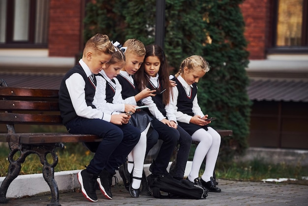 Ragazzi delle scuole in uniforme che siedono all'aperto in panchina con gli smartphone.