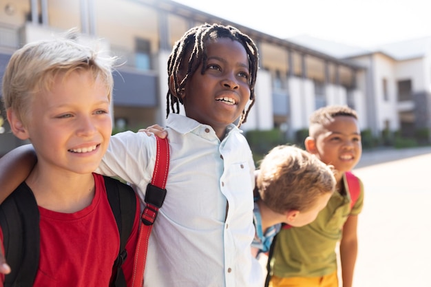 Ragazzi di scuola elementare multirazziali sorridenti con il braccio attorno in piedi nel campus della scuola durante una giornata di sole
