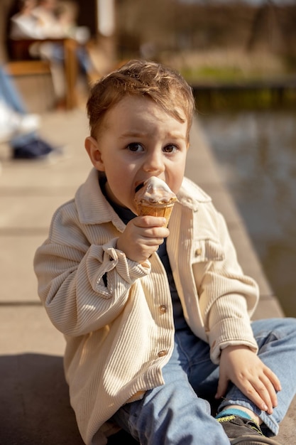 Ragazzino adorabile seduto all'aperto e mangiare gelato Acqua del lago e tempo soleggiato Bambino e dolci zucchero Il bambino si gode un delizioso dessert Bambino in età prescolare con abbigliamento casual Emozione positiva