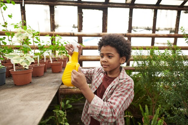 Ragazzino africano che tiene in mano una bottiglia e innaffia le piante verdi in giardino