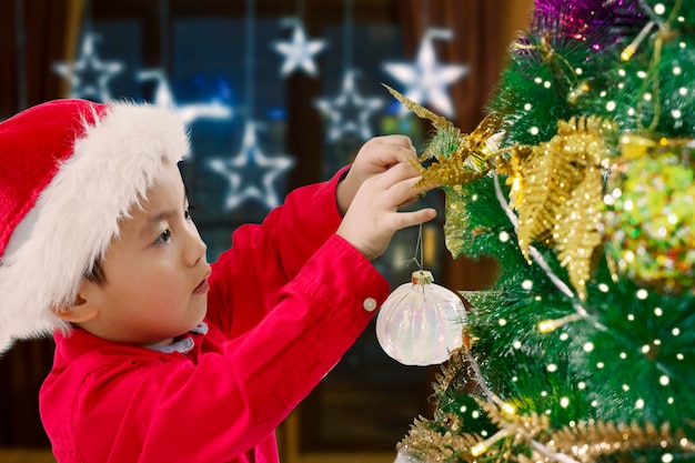 Ragazzino asiatico sveglio che appende una palla di Natale sull'albero di Natale a casa