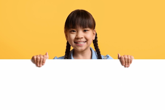 Ragazzino asiatico sveglio della ragazza che posa dietro il bordo di pubblicità in bianco