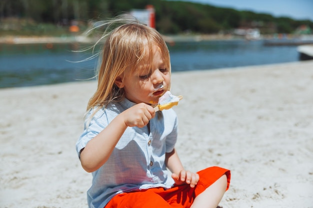 Ragazzino biondo carino con i capelli lunghi che mangia il gelato durante le vacanze estive sulla spiaggia
