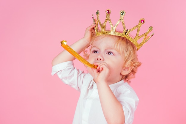 Ragazzino biondo con testa di corona dorata riccioli acconciatura 4-5 anni in studio su sfondo rosa che soffia noisemakers corno-fischio una festa di compleanno celebra il Natale e il nuovo anno re della festa