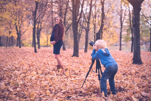 Ragazzino biondo con una grande macchina fotografica reflex su un treppiede. Fotografa una donna incinta. Sessione fotografica di famiglia