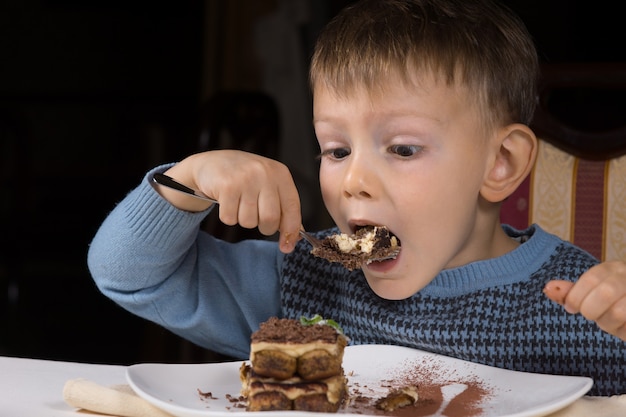 Ragazzino carino che mangia una torta al cioccolato che sta per prendere un grosso boccone con uno sguardo di impaziente attesa mentre si siede a tavola