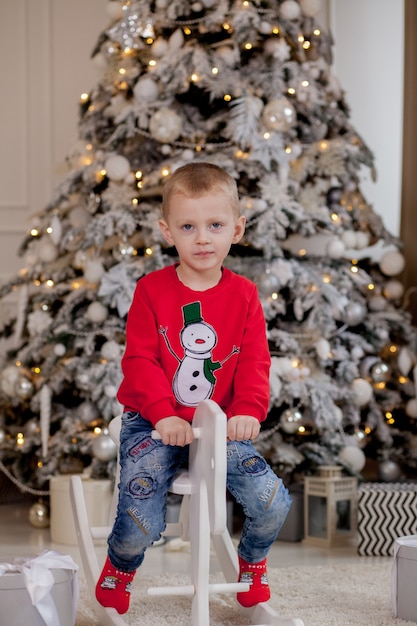 Ragazzino carino con accessori natalizi seduto su un cavallo di legno davanti all'albero di Natale