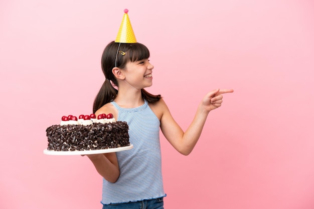 Ragazzino caucasico che tiene la torta di compleanno isolata su sfondo rosa che punta il dito di lato e presenta un prodotto