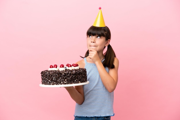 Ragazzino caucasico che tiene la torta di compleanno isolata su sfondo rosa con dubbi e pensieri