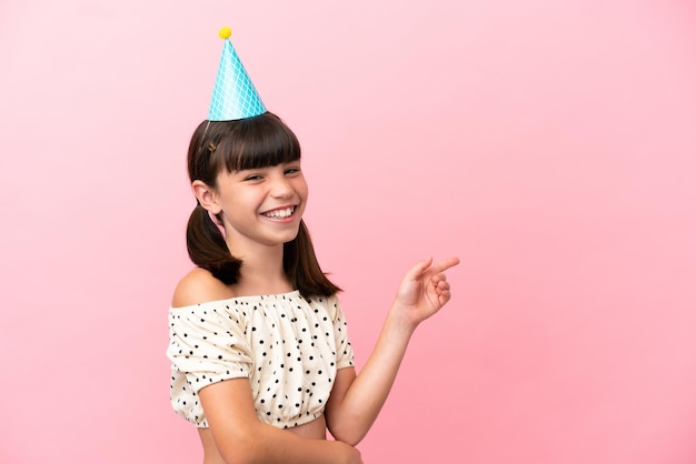 Ragazzino caucasico con cappello di compleanno isolato su sfondo rosa che punta il dito di lato