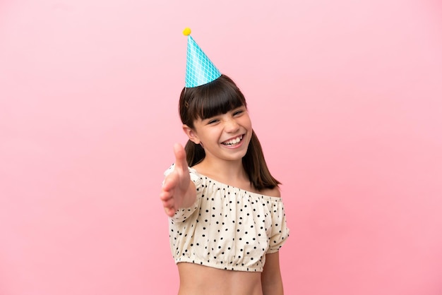 Ragazzino caucasico con cappello di compleanno isolato su sfondo rosa che stringe la mano per chiudere un buon affare