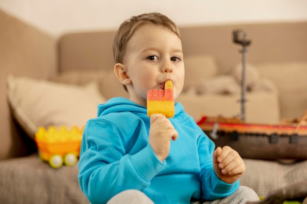 Ragazzino caucasico con felpa con cappuccio blu che gioca con i giocattoli colorati e mangia il gelato a casa Il bambino si diverte Il bambino felice e allegro gioca con il castello dei dinosauri della nave Attività per il tempo libero