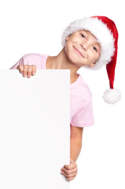 Ragazzino che indossa il cappello di Babbo Natale che posa dietro un pannello bianco isolato su uno sfondo bianco Bambino carino che tiene in mano un cartellone vacante di Natale Bambino che sbircia dietro un cartello vuoto di vacanze