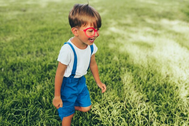 Ragazzino che indossa pantaloncini blu e occhiali rossi che giocano sullo sfondo della natura Bambino allegro che corre sull'erba verde sentendosi felice nel parco Bambino che si diverte all'aperto Infanzia felice