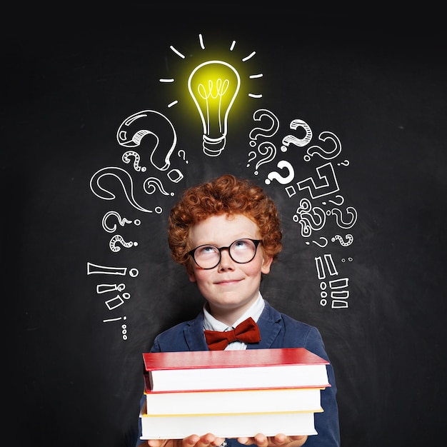 Ragazzino che indossa un papillon e un abito con una lampadina e libri sullo sfondo della lavagna Brainstorming e concetto di idea