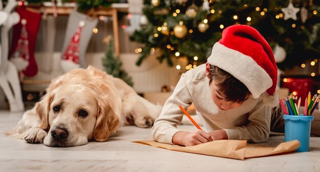 Ragazzino che scrive una lettera a Babbo Natale mentre giace accanto al cane golden retriever sul pavimento a casa