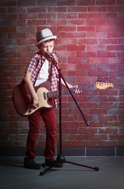 Ragazzino che suona la chitarra e canta con il microfono su uno sfondo di muro di mattoni