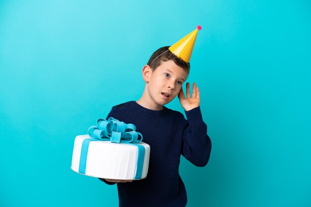 Ragazzino che tiene la torta di compleanno isolata su sfondo blu ascoltando qualcosa mettendo la mano sull'orecchio