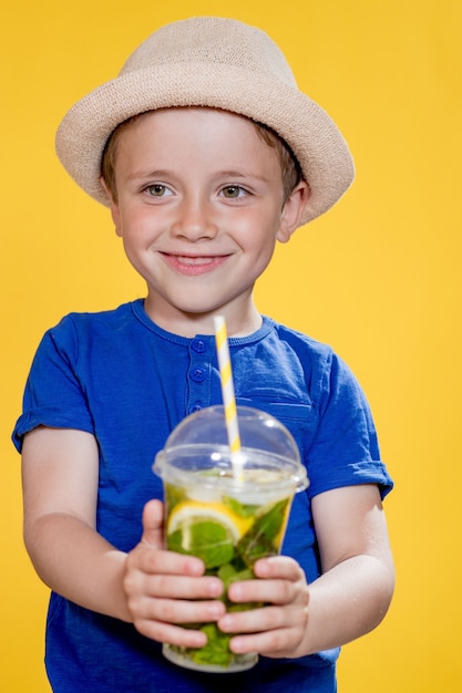 Ragazzino con cappello e maglietta blu che tiene cocktail mojito in una tazza di plastica su sfondo giallo