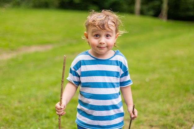 Ragazzino con i capelli sudati che tiene i bastoni e gioca nel parco nella stagione calda estiva.