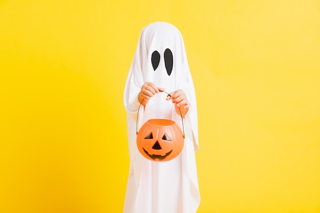 Ragazzino con il fantasma di halloween vestito bianco costume spaventoso che tiene il fantasma arancione della zucca a portata di mano