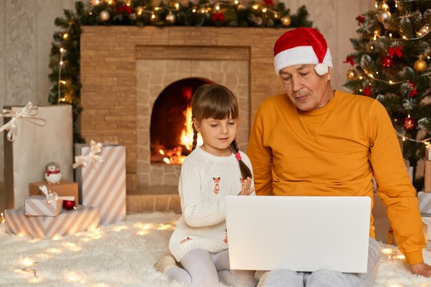 Ragazzino con il nonno seduto e utilizzando il notebook durante la mattina di Natale, guarda lo schermo, si siede sul pavimento vicino al camino in un accogliente soggiorno, uomo anziano in camicia gialla e cappello da Babbo Natale.