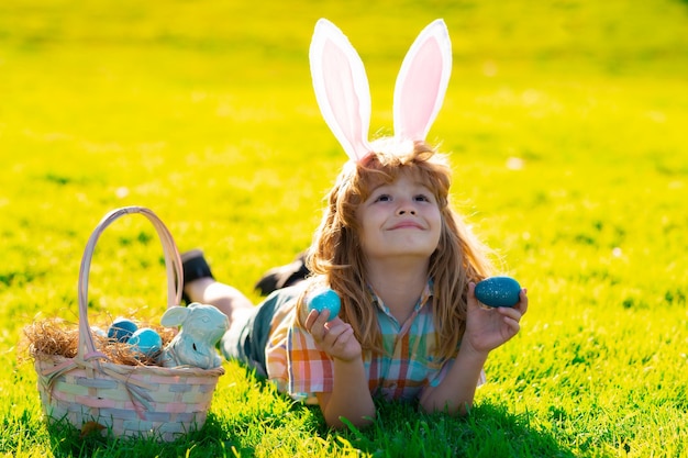 Ragazzino con orecchie da coniglio che cacciano le uova di Pasqua all'aperto Bambino che ha Pasqua posa sull'erba Caccia alle uova di Pasqua